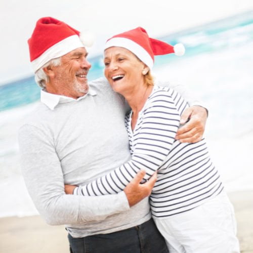 Older couple both wearing red santa hats embracing on the beach